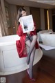 A woman in a red robe sitting on a bathtub holding a newspaper.